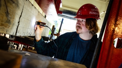 White male graduate student wearing a red hard had with the Rutgers logo holds a device to a beam in a construction lab. He has red hair and is wearing a green and navy blue flannel shirt.