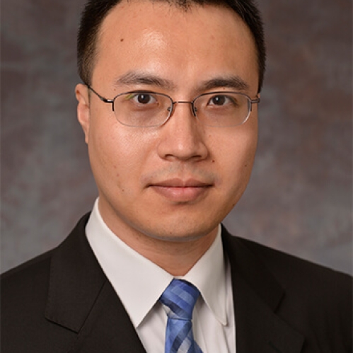 Asian male with eyeglasses wearing a dark suit, white shirt, and a blue patterned tie