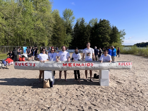 Group of students standing on sandy beach behind a grey concrete canoe. 