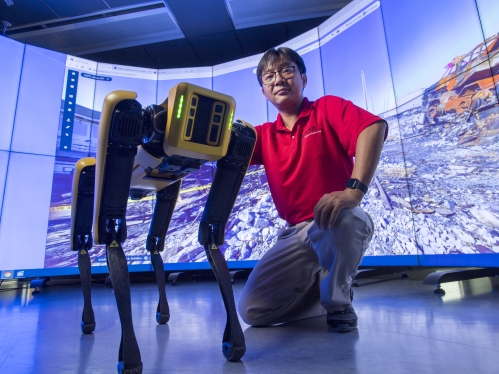 Asian male with black hair and glasses wearing a red polo shirt poses with a robotic yellow dog.