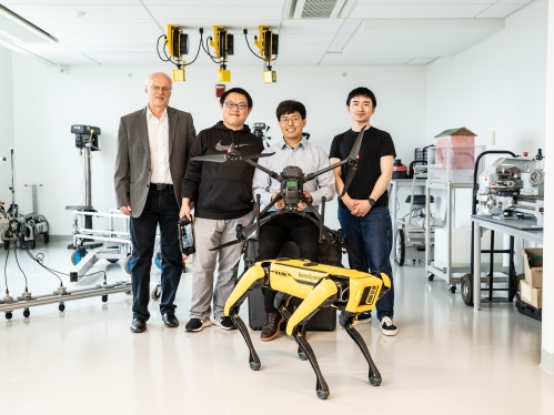 Four men pose in a lab with a yellow robotic dog and a drone.