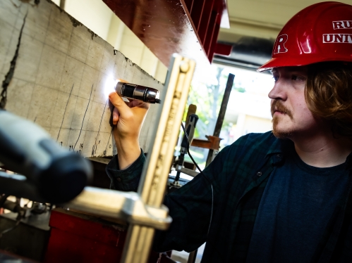 White male student in a red Rutgers hard hat holds a flashlight to a concrete beam in a lab setting.