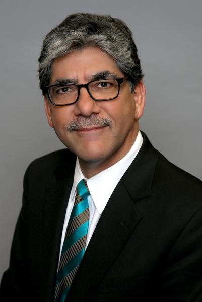 Headshot of man with salt and pepper hair, dark glasses, wearing a black suit, white shirt and green striped tie.