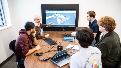 Professor points to a television monitor screen with a 3D buildig desgn as he instructs four students.