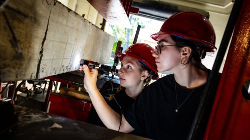 Two female students in red hard hats use a flashlight to exam concrete slab in a structural lab.