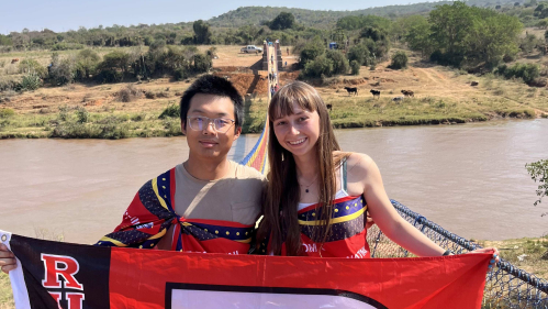 2 students stand on a bridge overlooking river, holding a Rutgers "R" flag together