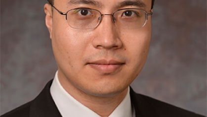 Asian male with eyeglasses wearing a dark suit, white shirt, and a blue patterned tie