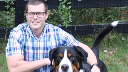 man with eyeglasses wearing a plaid shirt and jeans down on one knee with his arm around a medium size dog next to him 