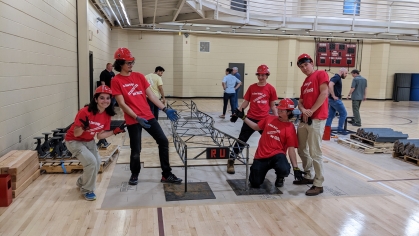 Five civil engineering students all wearing red Rutgers T-shirts and hard hats strike a pose around the steel bridge they constructed inside a gymnasium space.