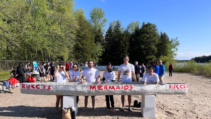 Civil engineering students pose with their concrete canoe on the sandy banks of a lake.