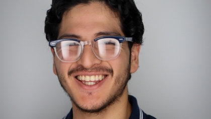Head shot of a white male college student with black framed glasses, black hair and a beard. He is wearing a navy blue polor shirt.