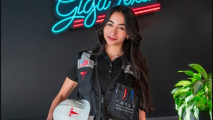Female engineering student poses wearing a work vest and holding under her arm a hard hat. She is standing in front of the neon sign that says Giga Texas. She has long black hair.