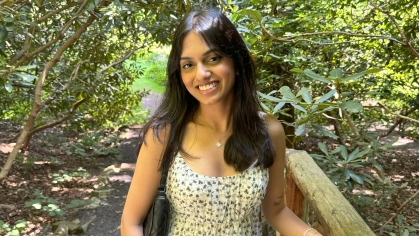 Student poses outdoors with trees in the background. She has long black hair and is wearing a yellow sundress.