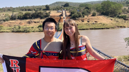 2 students stand on a bridge overlooking river, holding a Rutgers "R" flag together