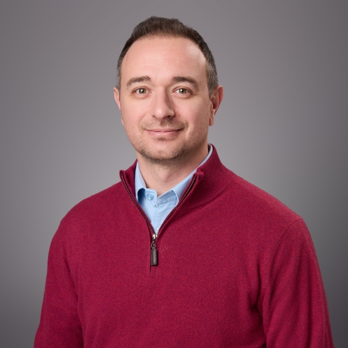 Headshot of male with short brown hair wearing a light blue shirt under a burgundy half zip sweater.