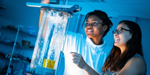 Two female students looking at a vessel holding water. The room is lit blue.