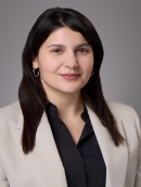 Headshot of woman with long brown hair wearing a black blouse and a light brown jacket.