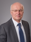 Headshot of male with grey hair and eyeglasses, wearing a grey suit, white shirt, and blue patterned tie.