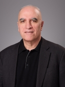 Headshot of male with short grey hair wearing a black button down shirt and a black suit jacket.