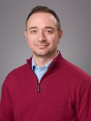 Headshot of male with short brown hair wearing a light blue shirt under a burgundy half zip sweater.