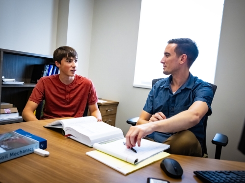 A professor meets with a student in his office. Sitting behind a desk the male professor is white with a blue short sleeved shirt, the male student is wearing a red short sleeved shirt. 