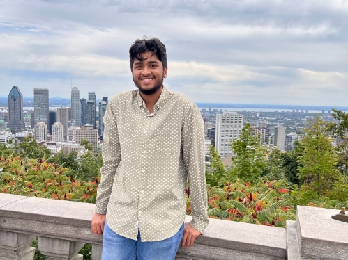 Male college student poses outdoors on a patio overlooking a city skyline. He is smiling wearing a light colored buttoned shirt and jeans.