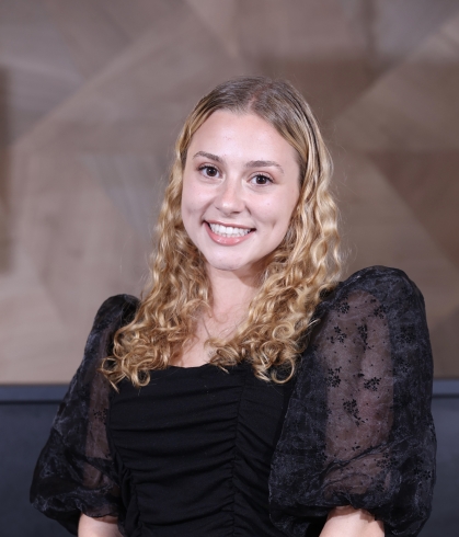 Young white woman with blond curly hair and brown eyes smiles at camera. She is wearing a black top with puffy sheer sleeves.