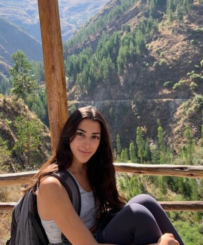 Young woman with long dark hair sitting with a back pack with a view of trees and mountains behind her.