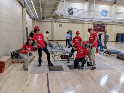 Five civil engineering students all wearing red Rutgers T-shirts and hard hats strike a pose around the steel bridge they constructed inside a gymnasium space.
