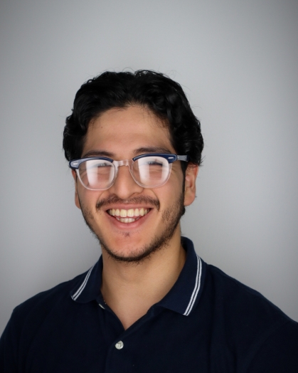 Head shot of a white male college student with black framed glasses, black hair and a beard. He is wearing a navy blue polor shirt.