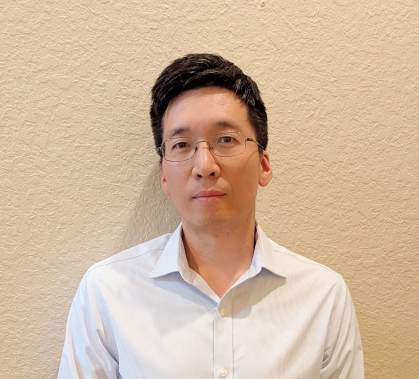 Headshot of man with black hair and glasses wearing an open collared white shirt.