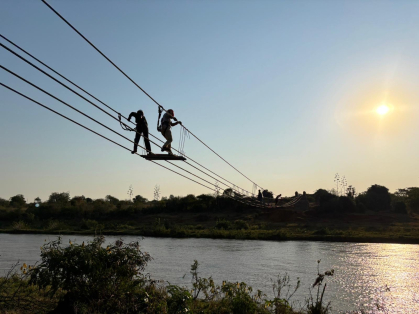 2 people standing on a platform tethered to a rope bridge in progress, with river underneath and sun against a soft blue sky visible