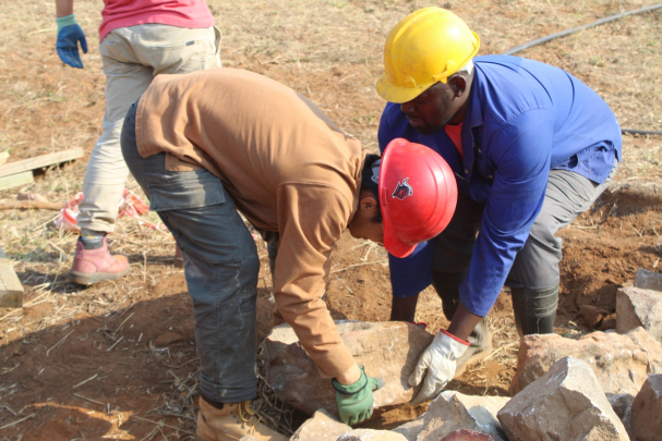 Student and worker stooping down to hold large stone together, with other stones nearby