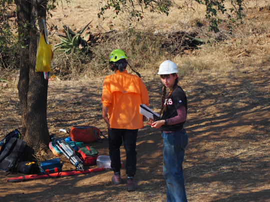 2 university students wearing hard hats stand under a tree, with various types of equipment laid on the dirt ground