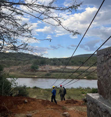 2 students stand in the grassy area on one side of a river, with ropes spanning the river hanging above them