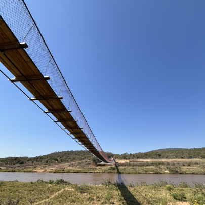 Full footbridge spanning the river is shown, with small hills in the background under a clear blue sky