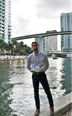 Full body photo of Ian Walczak standing on bank of river with tall building behind him
