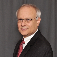 head shot of balding man with white hair and eyeglasses wearing a dark brown suit, white shirt, and maroon patterned tie