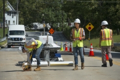 Three men wearing hard hats and yellow safety vests inspecting road with fully automated robotic device for making comprehensive condition assestment. 
