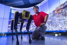 Asian male with black hair and glasses wearing a red polo shirt poses with a robotic yellow dog.