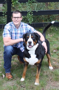 man with eyeglasses wearing a plaid shirt and jeans down on one knee with his arm around a medium size dog next to him 